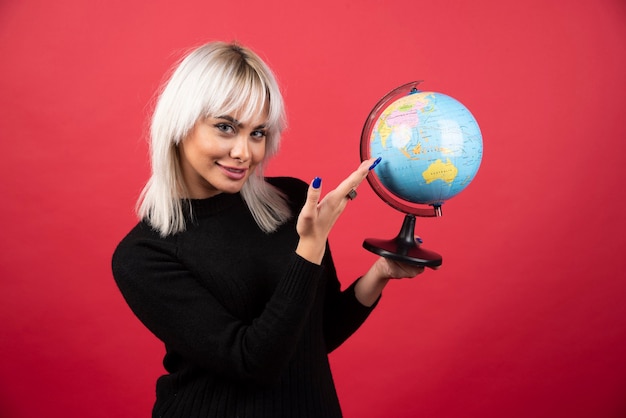 Junge Frau, die einen Globus auf einem roten Hintergrund zeigt. Hochwertiges Foto