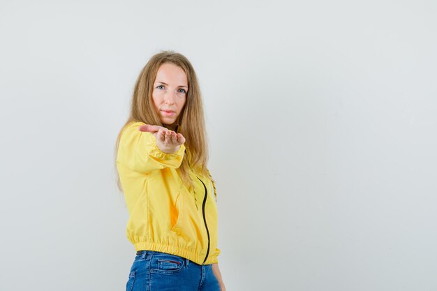 Junge Frau, die einlädt, in der gelben Bomberjacke und in der blauen Jeans zu kommen und ernst zu schauen. Vorderansicht.