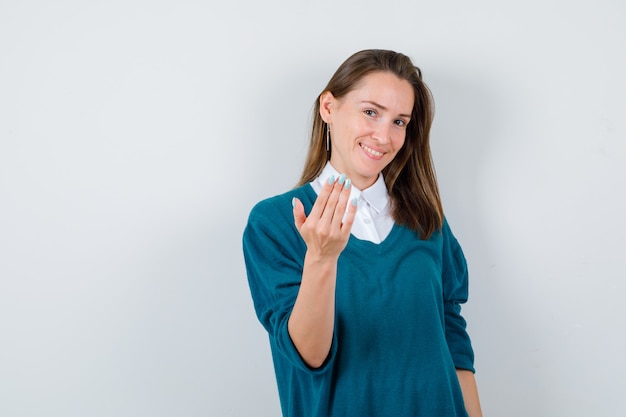 Junge Frau, die einlädt, in Pullover über weißem Hemd zu kommen und fröhlich zu schauen. Vorderansicht.