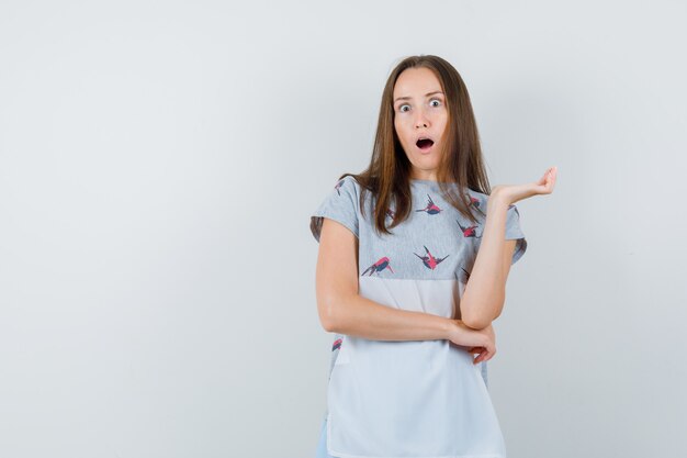 Junge Frau, die erhabene Handfläche im T-Shirt hält und überrascht, Vorderansicht schaut.