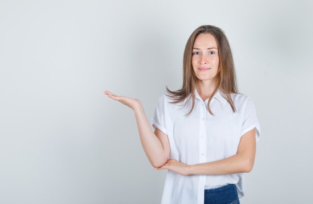 Junge Frau, die etwas im weißen T-Shirt, in den Jeans begrüßt oder zeigt und fröhlich aussieht