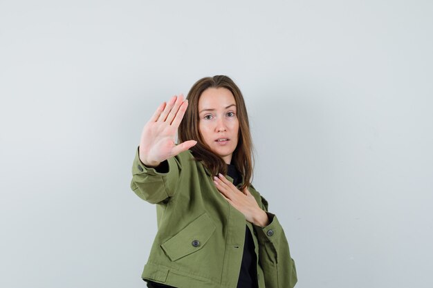 Junge Frau, die etwas mit Handbewegung in grüner Jacke ablehnt und vorsichtig schaut. Vorderansicht.