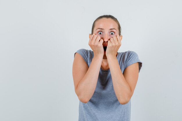 Junge Frau, die Fäuste auf Gesicht im grauen T-Shirt hält und ängstlich schaut. Vorderansicht.