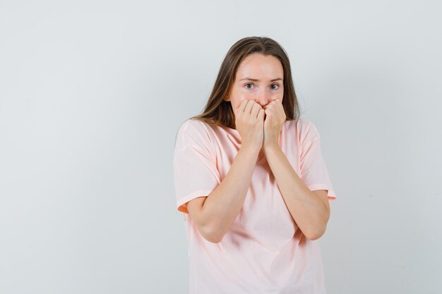 Junge Frau, die Fäuste auf Mund im rosa T-Shirt hält und ängstlich schaut. Vorderansicht.