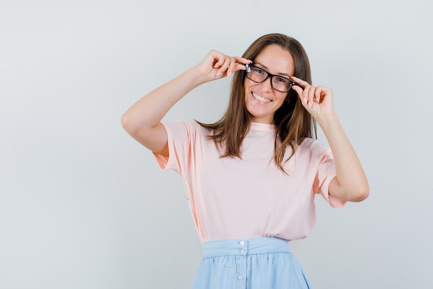 Junge Frau, die Finger auf Brille im T-Shirt, im Rock hält und fröhlich schaut. Vorderansicht.