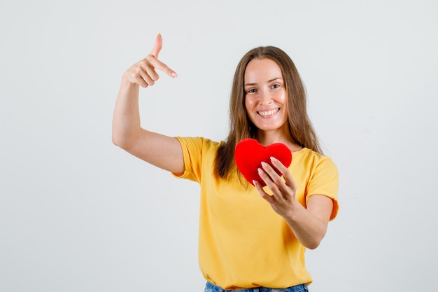 Junge Frau, die Finger auf rotes Herz im T-Shirt, in den Shorts zeigt und froh schaut