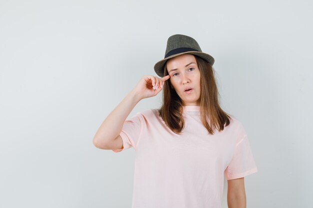 Junge Frau, die Finger auf Schläfen in rosa T-Shirt, Hut hält und vernünftig aussieht. Vorderansicht.