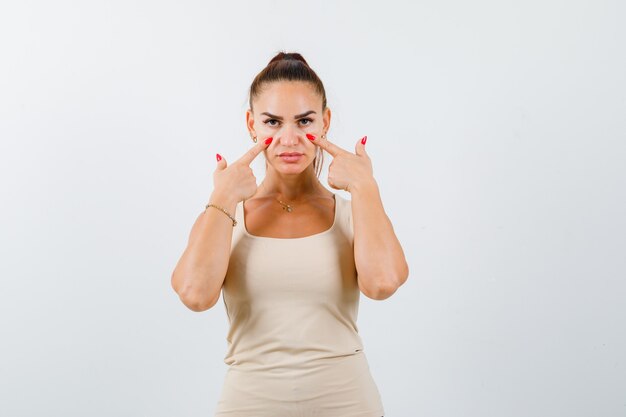 Junge Frau, die Finger auf Wangen im beigen Trägershirt hält und selbstbewusst, Vorderansicht schaut.