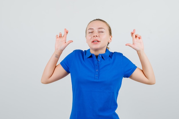 Junge Frau, die Finger im blauen T-Shirt gekreuzt hält und hoffnungsvoll aussieht