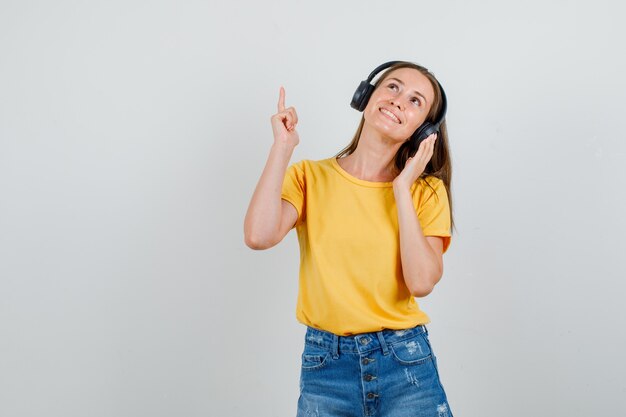 Junge Frau, die Finger oben in T-Shirt, Shorts, Kopfhörern zeigt und froh aussieht. Vorderansicht.