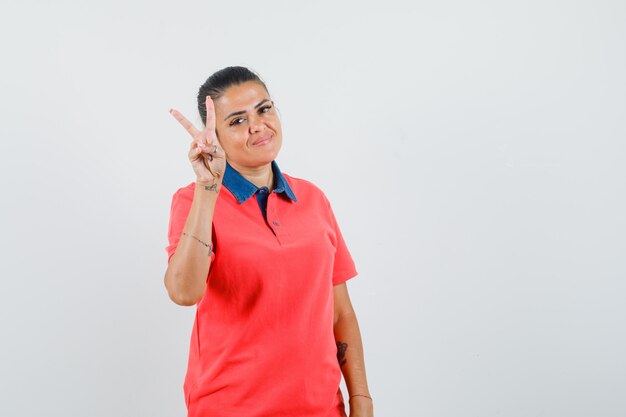 Junge Frau, die Friedenszeichen im roten T-Shirt zeigt und hübsch, Vorderansicht schaut.