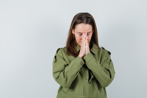 Junge Frau, die Gebetsgeste in der grünen Jacke zeigt und hoffnungsvoll aussieht. Vorderansicht.