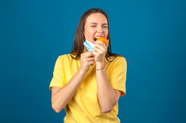 Junge Frau, die gelbes Poloshirt in der schützenden medizinischen Maske beißt orangefarbene Mandarine, die auf blauem Hintergrund steht