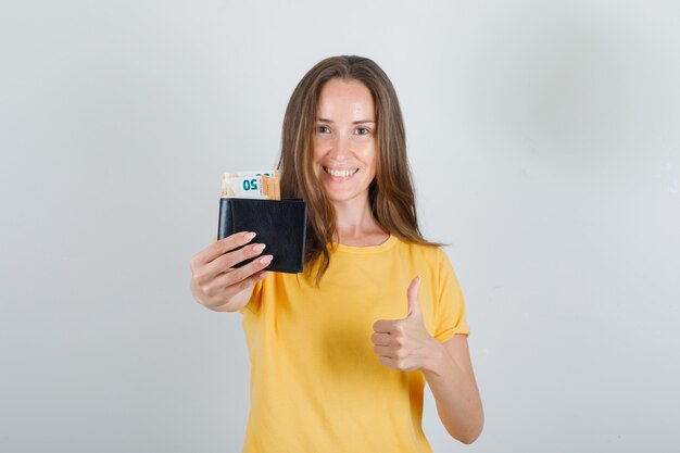 Junge Frau, die Geld in der Brieftasche mit Daumen oben im gelben T-Shirt hält und fröhlich schaut