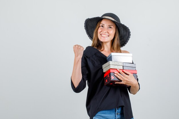 Junge Frau, die Geschenkboxen hält, während Gewinnerzeichen in Hemd, Shorts, Hut zeigt und fröhlich schaut. Vorderansicht.