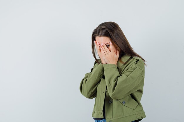 Junge Frau, die Gesicht mit Händen bedeckt, während durch Finger in grüner Jacke, Vorderansicht schaut. Platz für Text