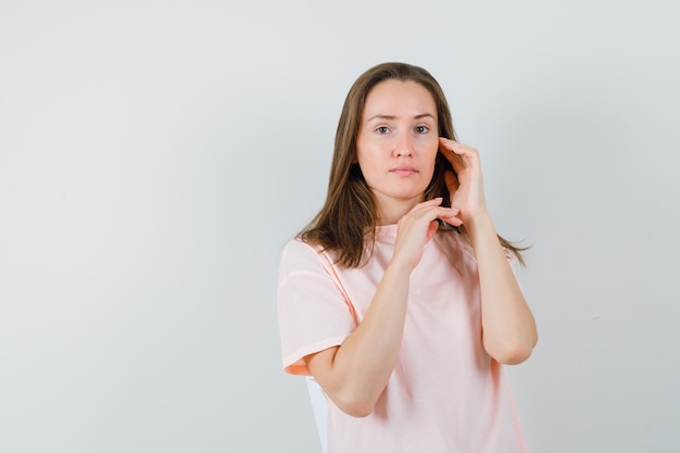 Junge Frau, die Gesichtshaut auf Wange im rosa T-Shirt berührt und zart schaut.