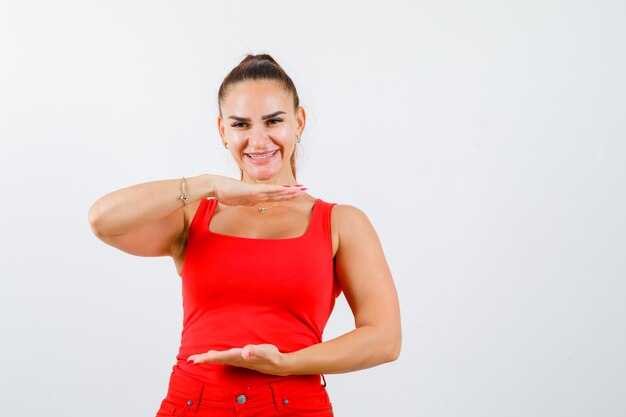 Junge Frau, die Größenzeichen im roten Trägershirt, in der Hose und im glücklichen Aussehen zeigt. Vorderansicht.