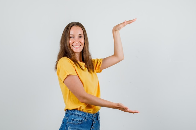 Junge Frau, die großes Zeichen in T-Shirt, Shorts zeigt und fröhlich schaut. Vorderansicht.