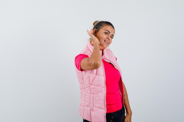Junge Frau, die Händchen in der Nähe des Ohres hält, um etwas in rosa T-Shirt und Jacke zu hören und neugierig zu schauen