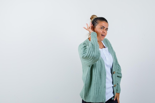 Junge Frau, die Händchen in der Nähe des Ohres hält, um etwas in weißem T-Shirt und mintgrüner Strickjacke zu hören und fokussiert zu schauen