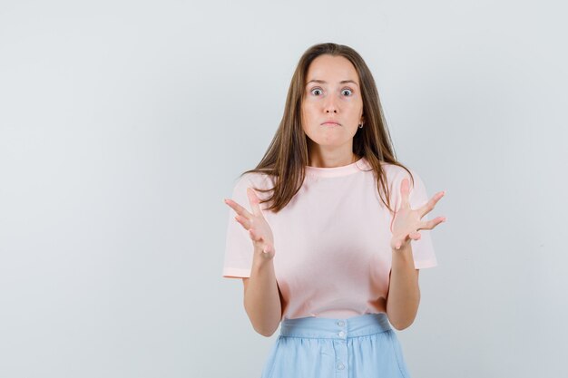 Junge Frau, die Hände auf aggressive Weise im T-Shirt, im Rock und im wütenden Blick erhebt. Vorderansicht.