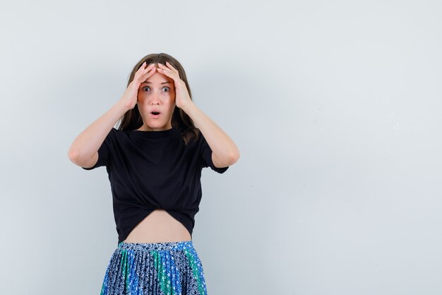 Junge Frau, die Hände auf Gesicht im schwarzen T-Shirt und im blauen Rock hält und überrascht schaut