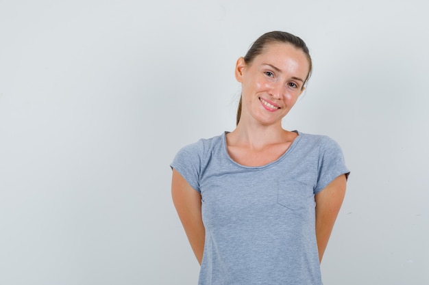 Junge Frau, die Hände auf zurück in grauem T-Shirt hält und fröhlich schaut. Vorderansicht.