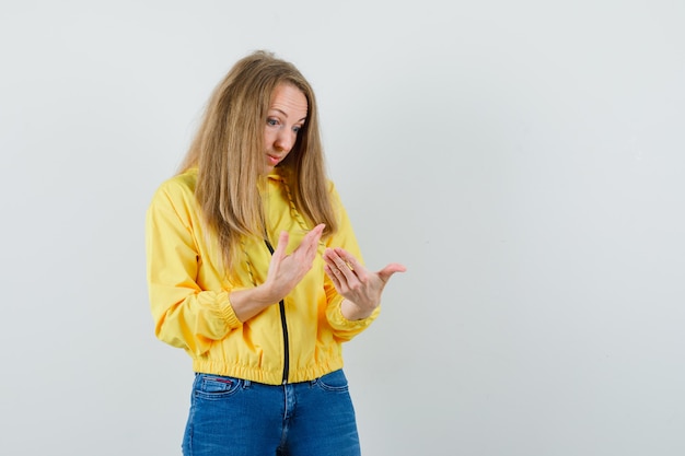 Junge Frau, die Hände betrachtet, wie sie etwas Imaginäres in gelber Bomberjacke und blauer Jeans hält und überrascht schaut. Vorderansicht.