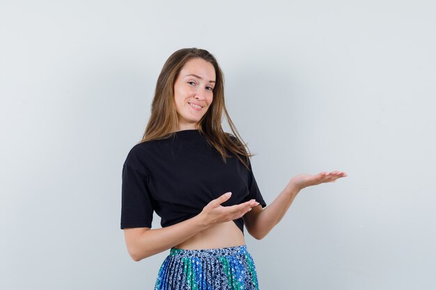 Junge Frau, die Hände hält, als etwas im schwarzen T-Shirt und im blauen Rock darstellend und glücklich aussehend