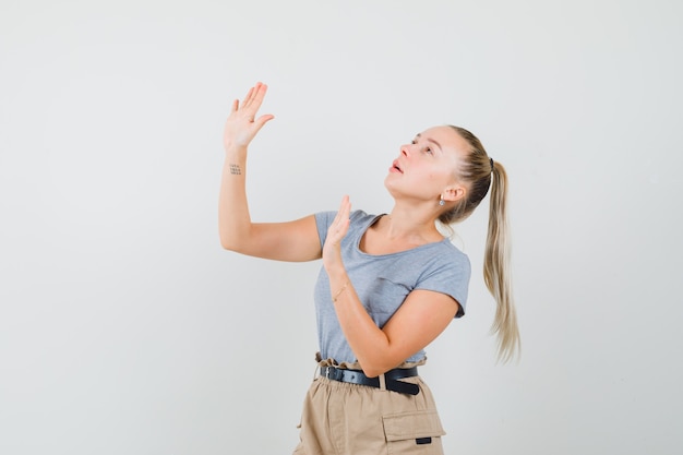 Junge Frau, die Hände hebt, um sich im T-Shirt, in der Hose und in der ängstlichen Vorderansicht zu verteidigen.