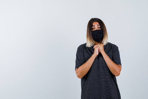 Junge Frau, die Hände in betender Position im schwarzen Kleid, in der schwarzen Maske und in der fokussierten Vorderansicht fasst.