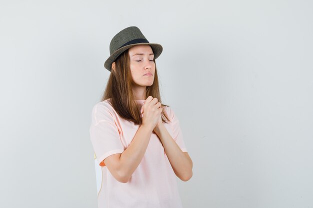 Junge Frau, die Hände in der Gebetsgeste im rosa T-Shirt, im Hut und im ruhigen Blick fasst. Vorderansicht.