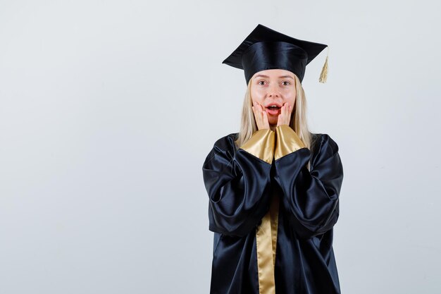 Junge Frau, die Hände in der Nähe des offenen Mundes in der Absolventenuniform hält und überrascht schaut