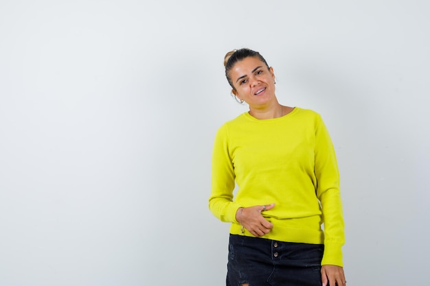 Junge Frau, die Hand auf Bauch in Pullover, Jeansrock hält und fröhlich aussieht looking