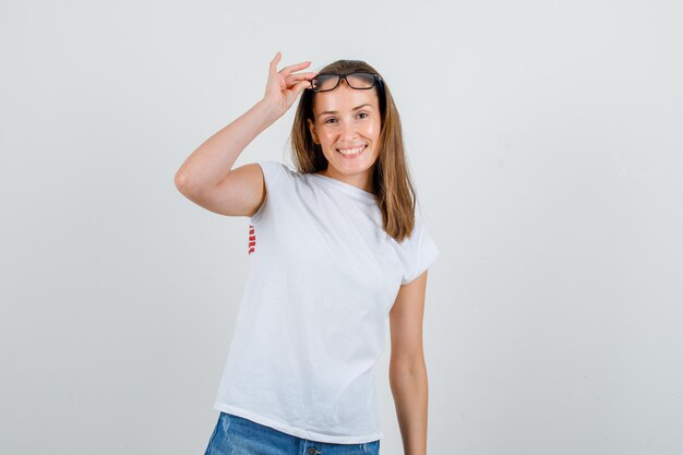 Junge Frau, die Hand auf Brille im T-Shirt, in den Shorts hält und fröhlich schaut. Vorderansicht.