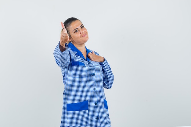 Junge Frau, die Hand auf Brust im blauen Gingham-Pyjamahemd zeigt und hält und hübsch, Vorderansicht schaut.