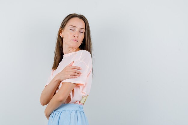 Junge Frau, die Hand auf dem anderen Arm in T-Shirt, Rock und friedlich aussehend hält. Vorderansicht.