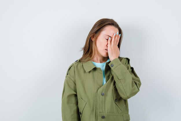 Junge Frau, die Hand auf Gesicht in grüner Jacke hält und deprimiert, Vorderansicht schaut.