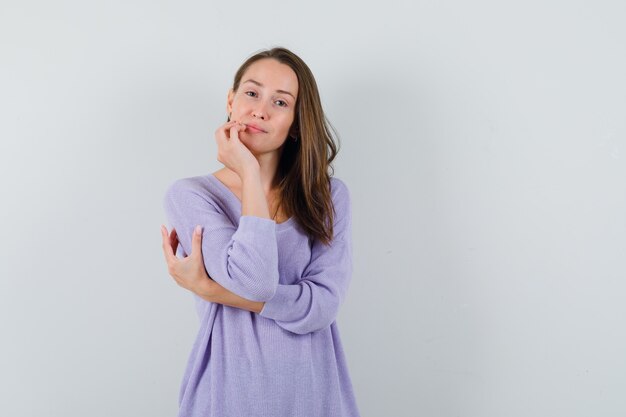 Junge Frau, die Hand auf ihrem Kinn hält, während sie ihren Ellbogen in der lila Bluse umarmt und beeindruckend aussieht