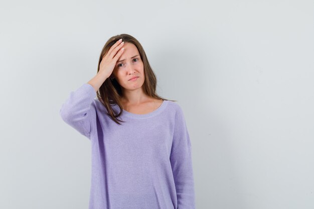 Junge Frau, die Hand auf ihrem Kopf in der lila Bluse hält und unangenehm aussieht. Vorderansicht.