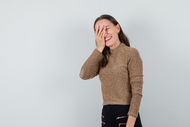 Junge Frau, die Hand auf ihren Augen in goldener Bluse hält und fröhlich schaut. Vorderansicht. Platz für Text