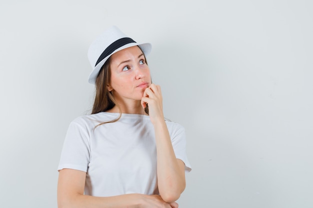 Junge frau, die hand auf kinn im weißen t-shirt, hut hält und nachdenklich schaut.