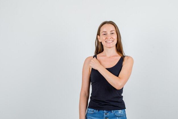 Junge Frau, die Hand auf Kleidern im schwarzen Unterhemd, in den kurzen Hosen und im fröhlichen Blick hält