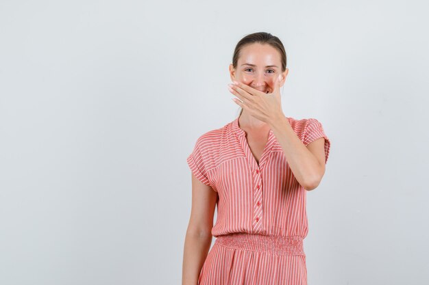 Junge Frau, die Hand auf Mund im gestreiften Kleid hält und glücklich schaut. Vorderansicht.