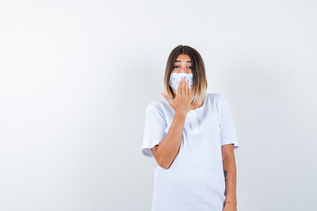 Junge Frau, die Hand auf Mund im T-Shirt, in der Maske hält und fröhlich, Vorderansicht schaut.