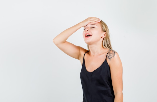 Junge frau, die hand auf stirn im schwarzen unterhemd hält und positiv schaut.