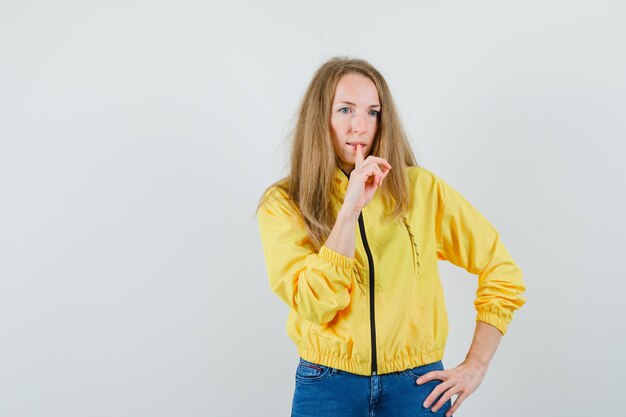 Junge Frau, die Hand auf Taille hält und Stille Geste in der gelben Bomberjacke und in der blauen Jeans zeigt und ernst schaut. Vorderansicht.