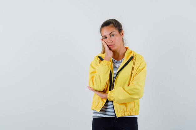 Junge Frau, die Hand auf Wange in Jacke, T-Shirt hält und hübsch, Vorderansicht schaut.