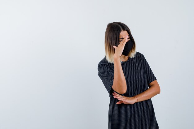 Junge Frau, die Hand erhebt, um Gesicht in schwarzem Kleid, schwarzer Maske zu verbergen und beschämt, Vorderansicht schauend.